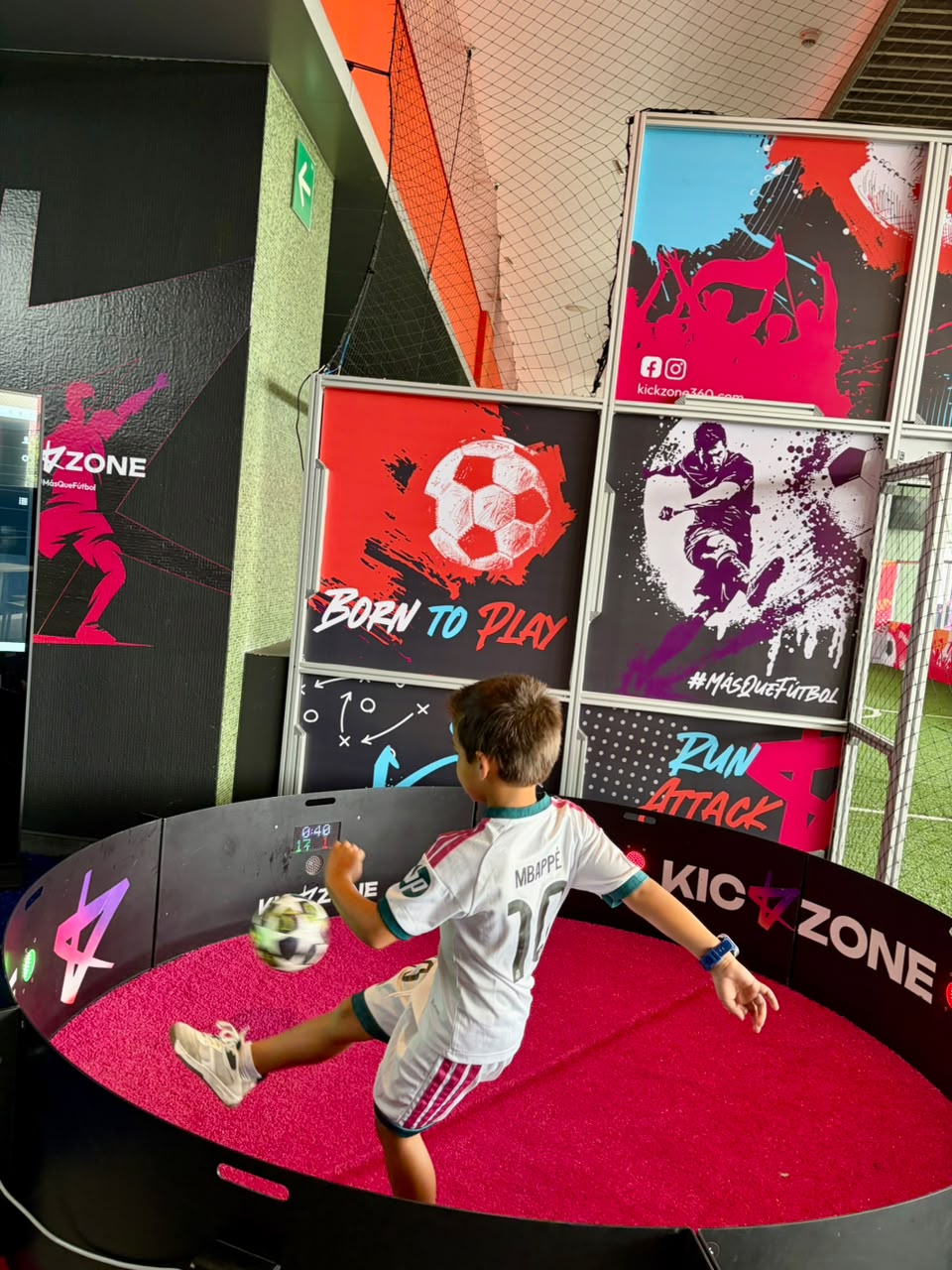 Niño con balón en el equipo circular de Kick Zone, con luces y pantalla de resultados
