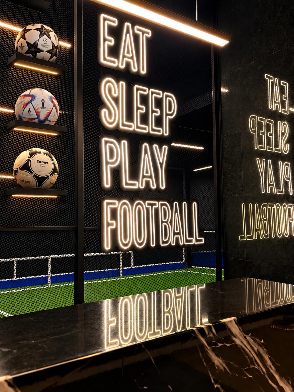 Recepción de Kick Zone con letrero luminoso Eat Sleep Play Football, balones iluminados y la cancha al fondo